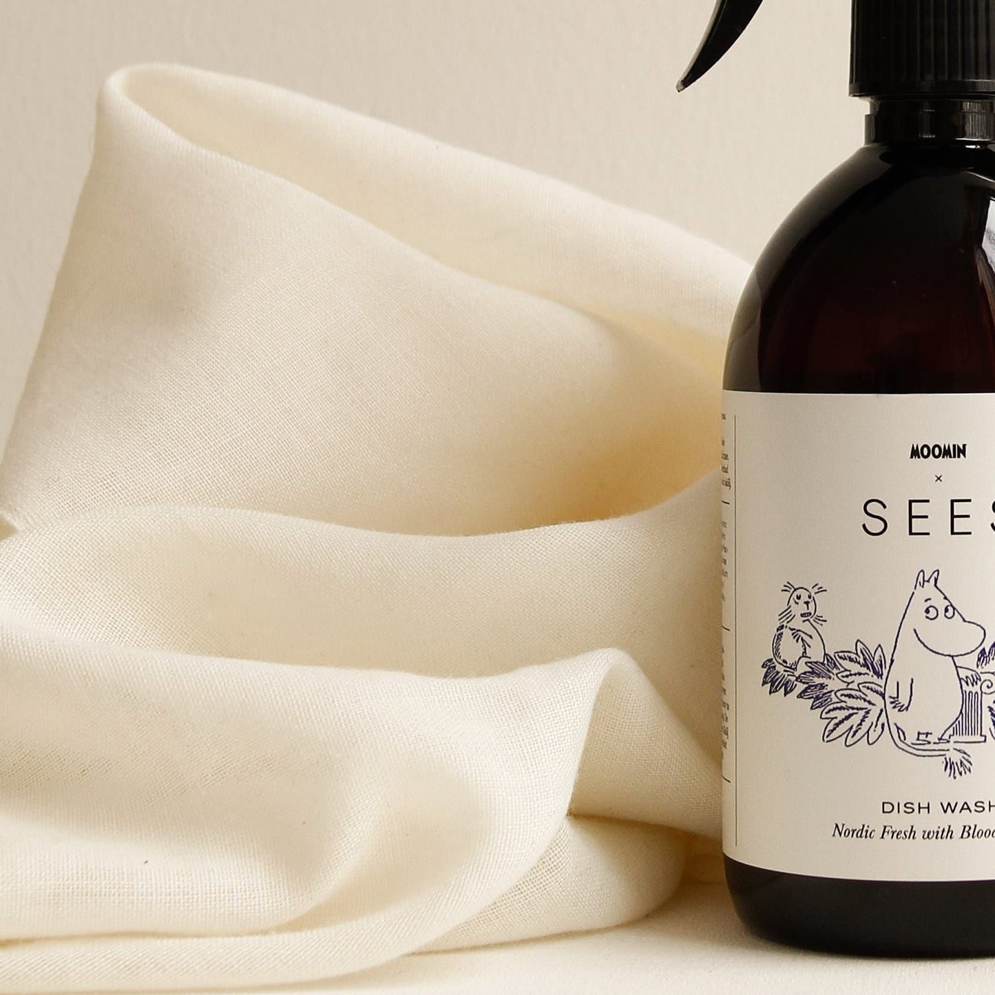 A SEES x Moomin Dish Wash Spray by SEES Company, featuring a brown bottle with a Moomin-themed label, stands beside a soft white cloth on a light, neutral background.