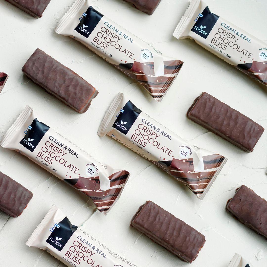 Foodin Clean & Real Protein Bar Crispy Chocolate Bliss bars and wrappers are laid out diagonally on a light surface. Each bar from Foodin is high in protein and contains no added sugar, making it a wholesome treat.