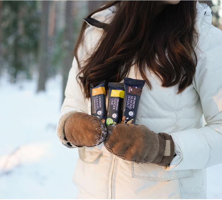 A person in a white winter jacket and brown mittens stands in a snowy forest, holding three Foodin Collagen Protein Bar Crunchy Almond Seasalt bars against their chest. Only the lower part of their face and hair are visible.
