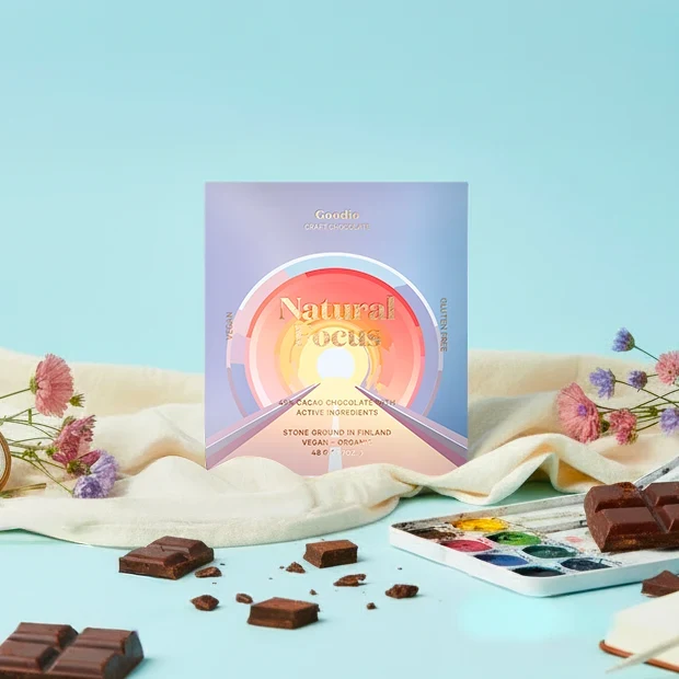A square chocolate gift box labeled 'Natural Focus' stands on a light blue surface with scattered chocolate pieces, cream fabric, small flowers, and a watercolor palette. The box has a pastel gradient cover with a circular sun design and text reading 'Goodie Chocolate Company' and 'Vegan Chocolate'.
