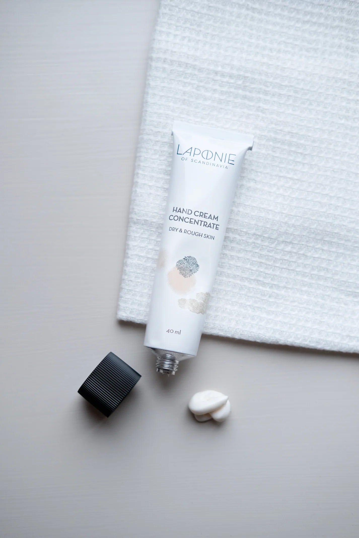 A tube of Laponie of Scandinavia Hand Cream Concentrate, cap off, sits on a light surface with some cream beside it and a textured white cloth in the background.