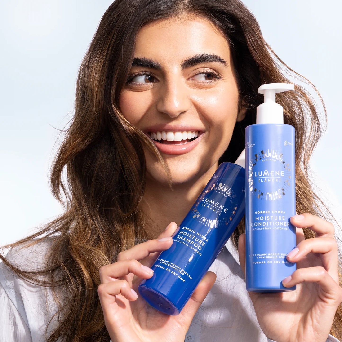 A smiling woman with long brown hair holds up two blue bottles of Lumene Nordic Hydra Moisture Shampoo against a light background.