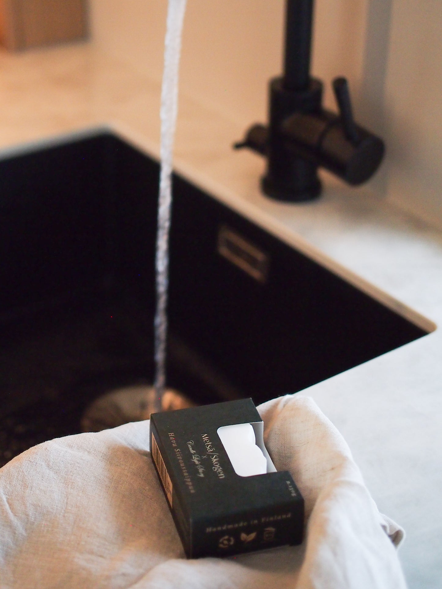 The Metsä/Skogen Conifer Cleaning Soap Bar by Metsä/Skogen, in a black box, sits on a beige cloth near a kitchen sink with water flowing from a black faucet, emphasizing its natural and eco-friendly cleaning appeal in a modern setting.