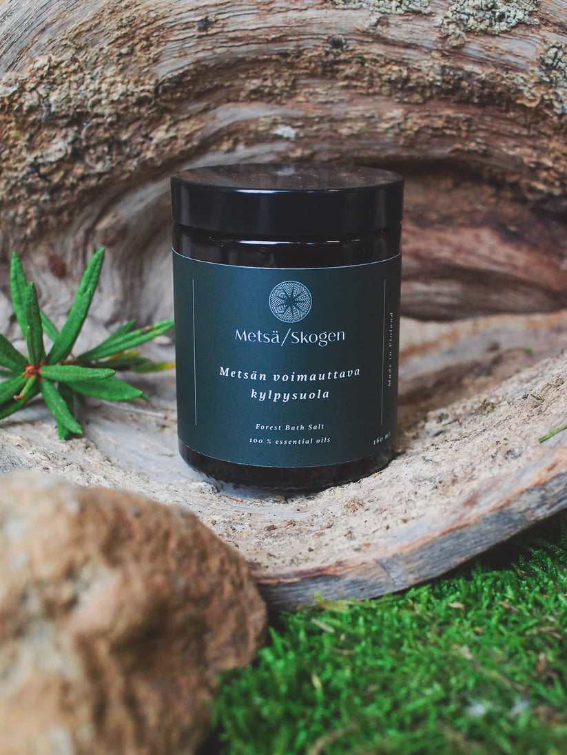 Metsä/Skogen Forest Bath Salt by Metsä/Skogen rests on moss with rocks, a curved wood piece, and fresh greenery nearby, evoking a soothing, natural forest-inspired aroma.