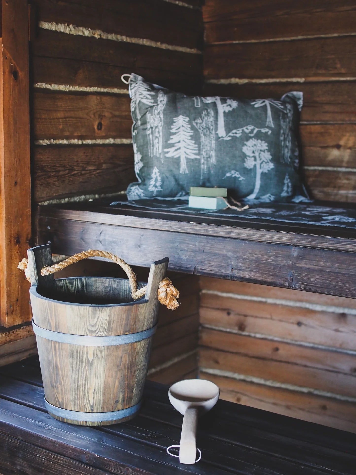 A rustic wooden sauna with a bench, a decorative tree-pattern pillow, soap, and a bucket with rope handle and wood ladle is filled with the calming Finnish forest aroma of Metsä/Skogen Forest Scent Drop by Metsä/Skogen.