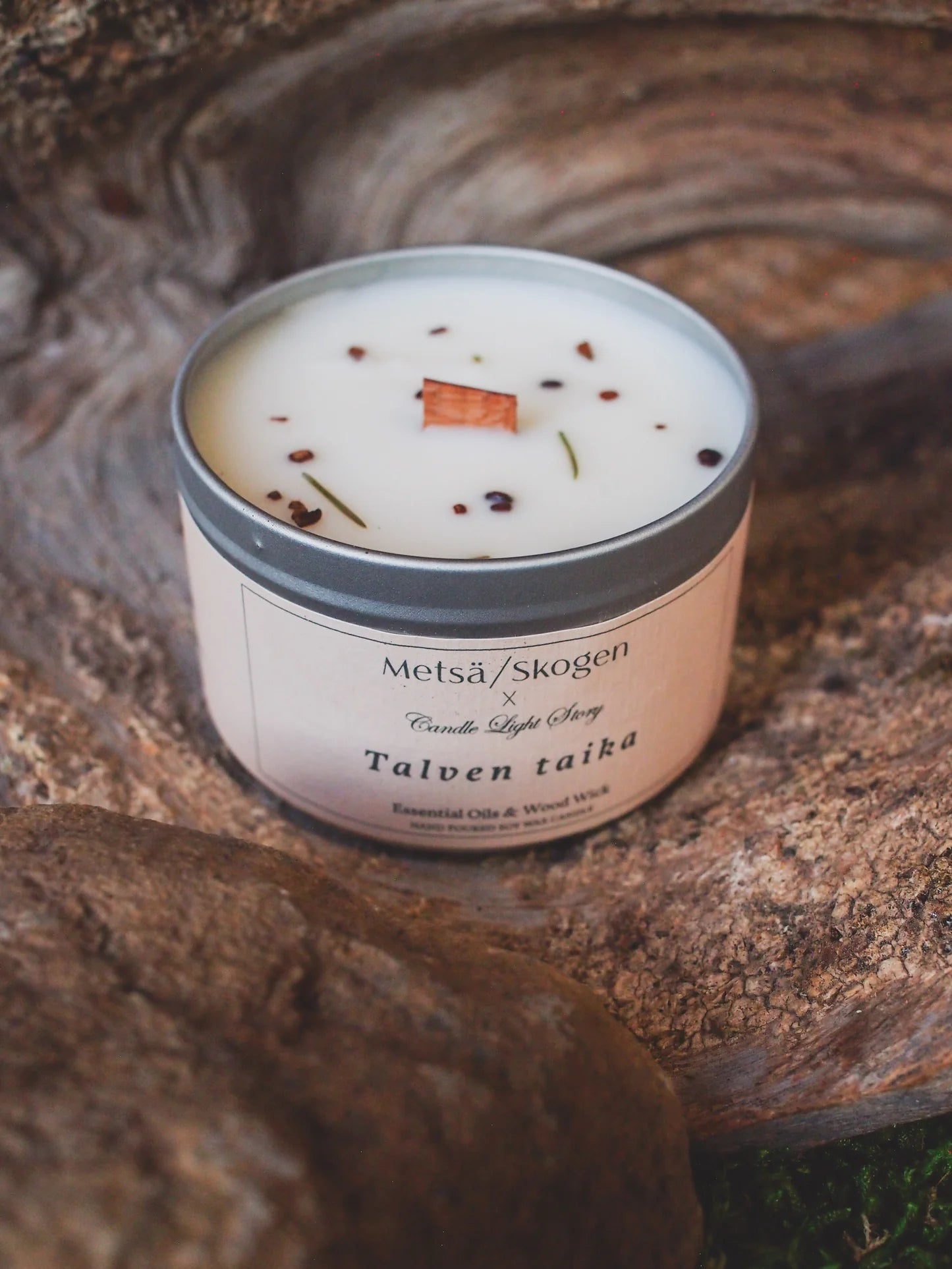 The Metsä/Skogen Winter’s Magic Soy Wax Candle sits in a white tin labeled with the brand, topped with a cinnamon stick, herbs, and spices—evoking Finnish forests and pine needles on textured wood.
