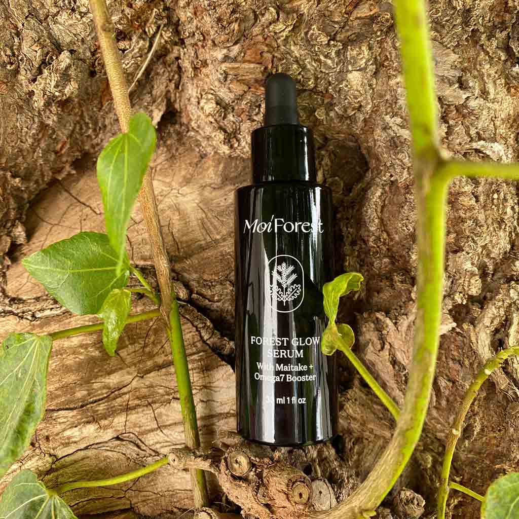 A black bottle of MoiForest Forest Glow Serum placed on textured tree bark with green leaves and vines around it.