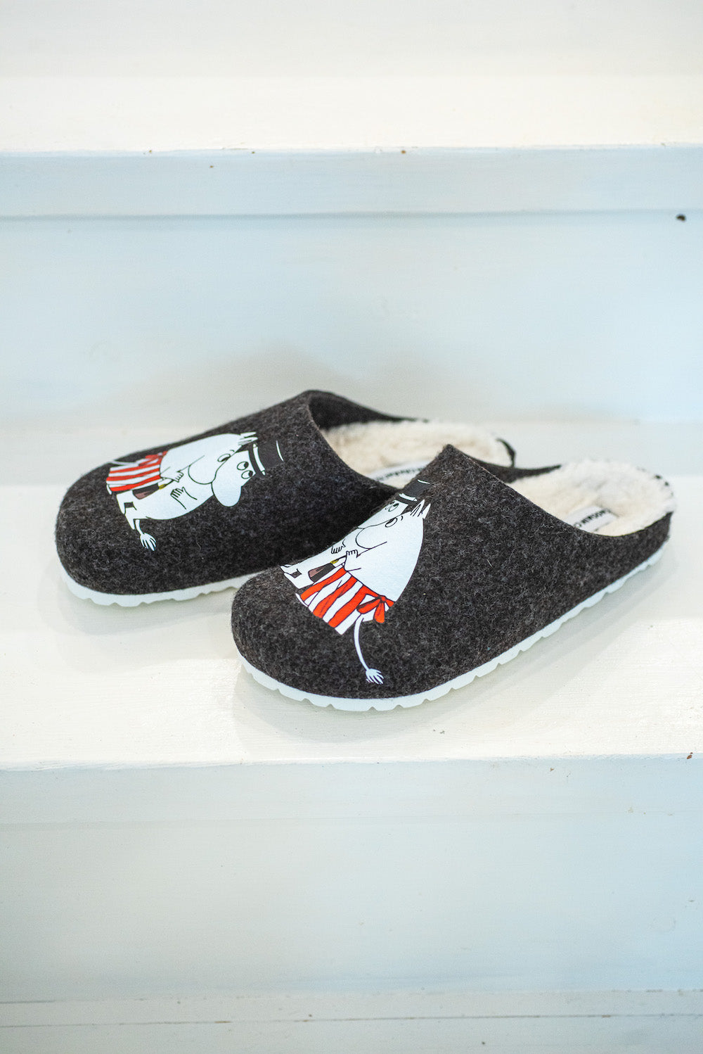 The Zapatillas Moomin Felt Slippers Moominmamma & Moominpappa are dark gray vegan slippers with a white furry lining and cartoon prints, shown on white wooden steps.