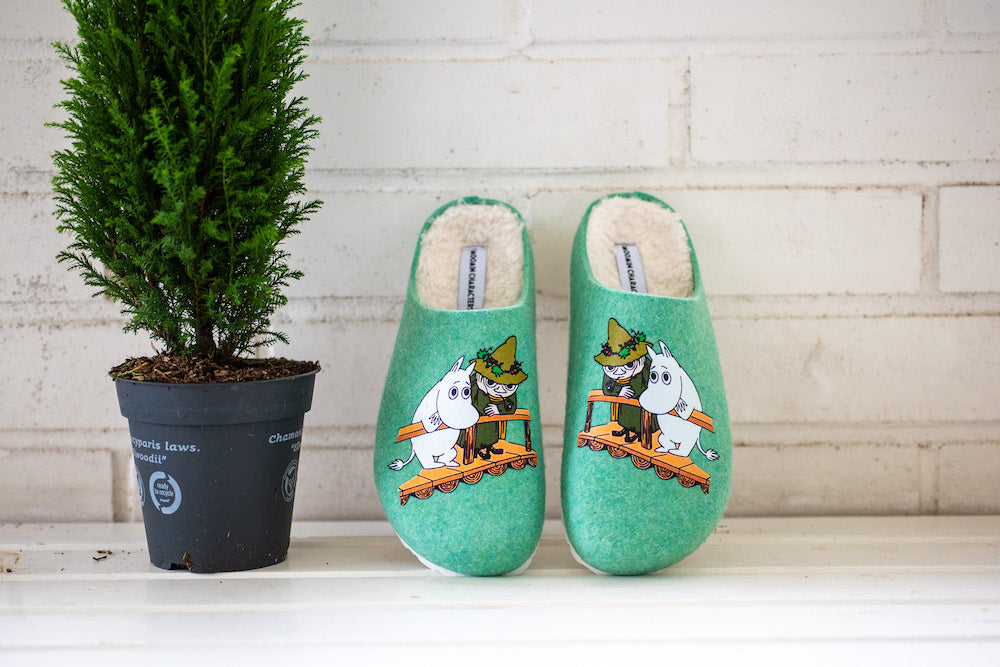 A pair of green Zapatillas Moomin Felt Slippers Snufkin with a charming character design rest by a small potted evergreen on a white shelf against a white brick wall, bringing cozy, eco-friendly comfort to your home.