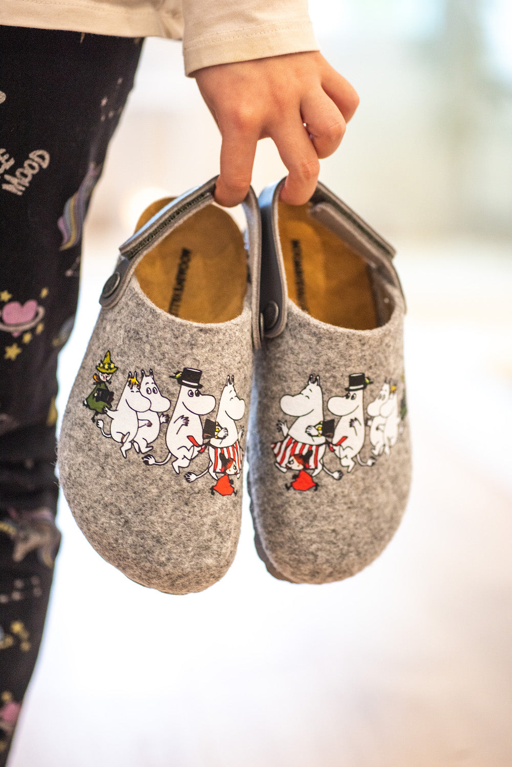 Someone holds a pair of Zapatillas Moomin Kids Felt Slippers Grey—cozy slippers featuring beloved characters—while wearing black patterned pants. The softly blurred background highlights the product.