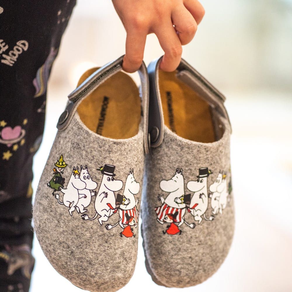 Someone holds a pair of Zapatillas Moomin Kids Felt Slippers Grey—cozy slippers featuring beloved characters—while wearing black patterned pants. The softly blurred background highlights the product.