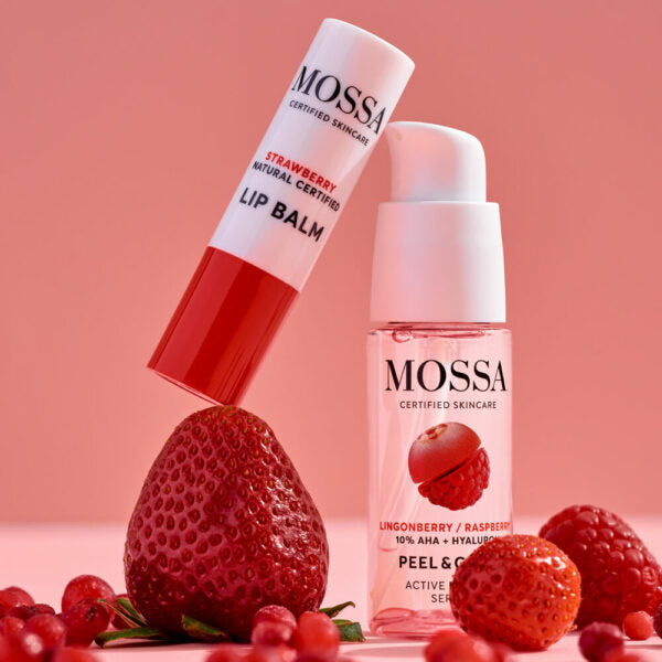 The Mossa Berry Kiss Gift Set featuring hydrating lip balm and overnight peeling serum is displayed among strawberries, raspberries, and lingonberries against a pink background.