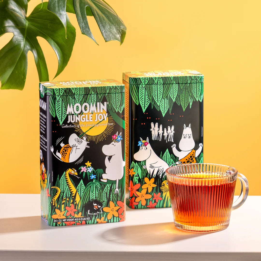 Two boxes of Moomin Jungle Joy tea with illustrated packaging, displayed on a white surface next to a glass mug filled with amber tea, against a yellow background with a green leaf in the corner.