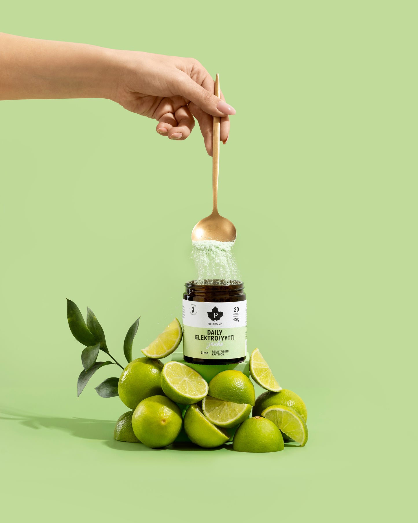 A hand holds a golden spoon with Puhdistamo Daily Electrolyte Powder Lime above a jar, surrounded by fresh whole and sliced limes, set against a light green background. The product is from the brand Puhdistamo.