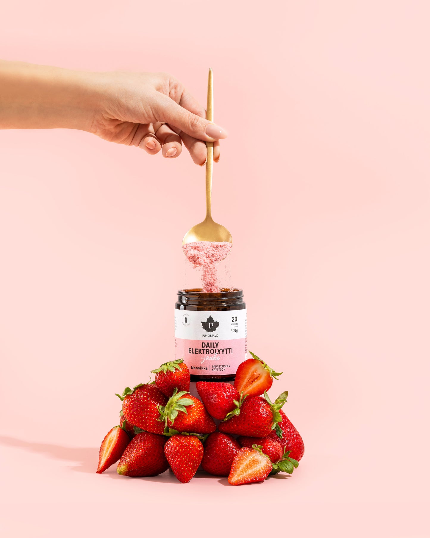 A hand holds a golden spoon above a jar of Puhdistamo Daily Electrolyte Powder Strawberry, surrounded by fresh strawberries against a pale pink background—showcasing daily hydration and natural strawberry flavor.