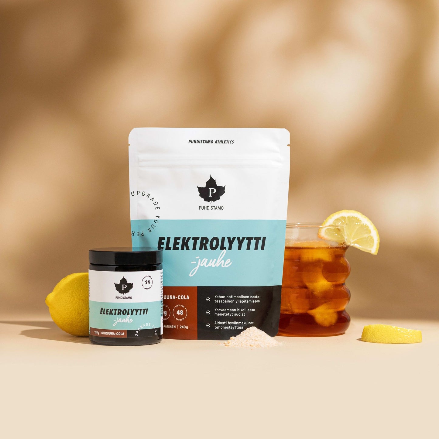 Puhdistamo Electrolyte Powder Lemon & Cola is shown with a jar, a scoop of powder, a glass of iced tea with lemon wedge, and lemon slices on a tan surface against a soft brown background—perfect for hydration and keto lifestyles.