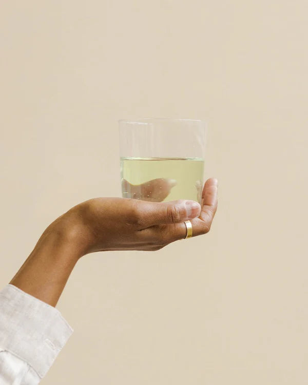 A hand with a gold ring holds up a clear glass of water mixed with Puhdistamo Electrolyte Powder Pear, from Puhdistamo, perfect for hydration. The person in a white long-sleeve shirt poses against a plain beige background.