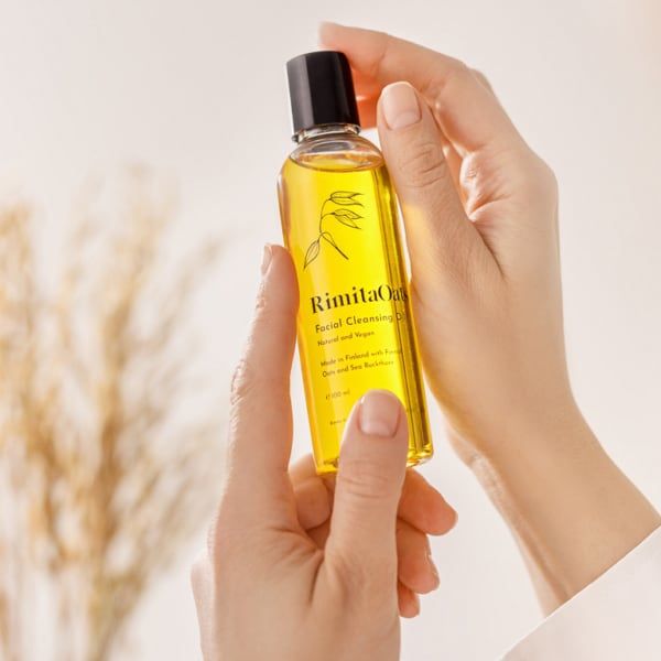 A person holds a clear bottle of RimitaOats Facial Cleansing Oil by Rimita Green, suitable for dry sensitive skin, with a black cap. Blurred dried plants appear in the softly lit background.