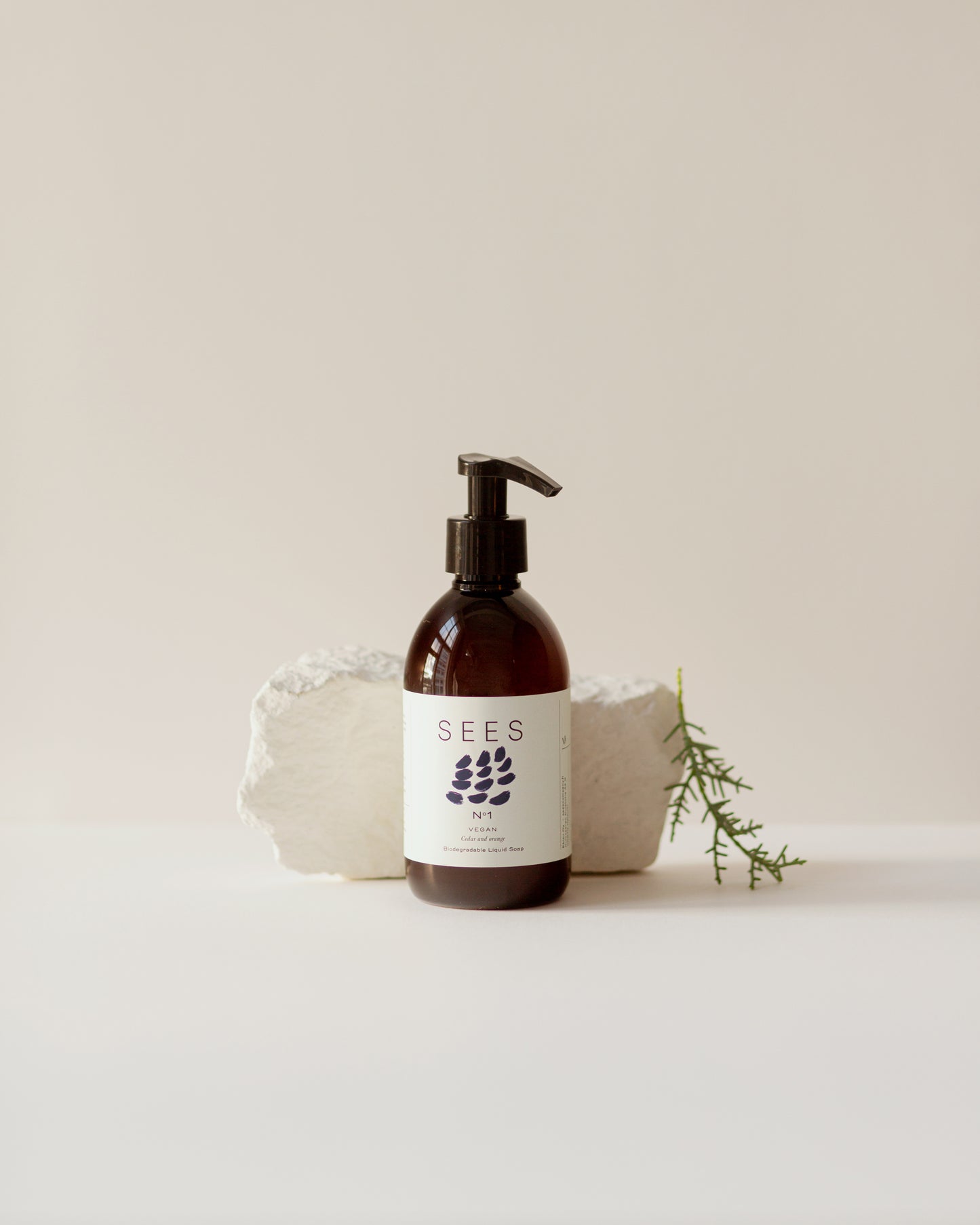 A brown pump bottle of SEES Company’s Hand Wash No. 1 Cedar & Orange stands before two white textured stones and a green sprig, set against a neutral background.