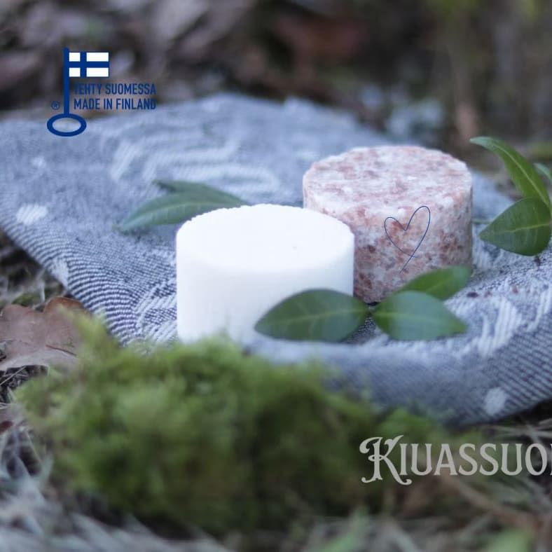Zwei runde Kiuassuola-Sauna-Salzseifen, eine weiße und eine rosa gesprenkelte, ruhen auf einem gemusterten Tuch im Freien inmitten von grünen Blättern und Moos. Ein "Made in Finland"-Etikett unterstreicht ihre Herkunft und ihre Verbindung zur traditionellen finnischen Saunapflege.