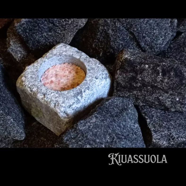 The Kiuassuola Sauna Salt ILMARI Starter Pack features a pinkish natural sauna salt block in a square stone container, surrounded by dark stones—perfect for enhancing your wellness ritual.