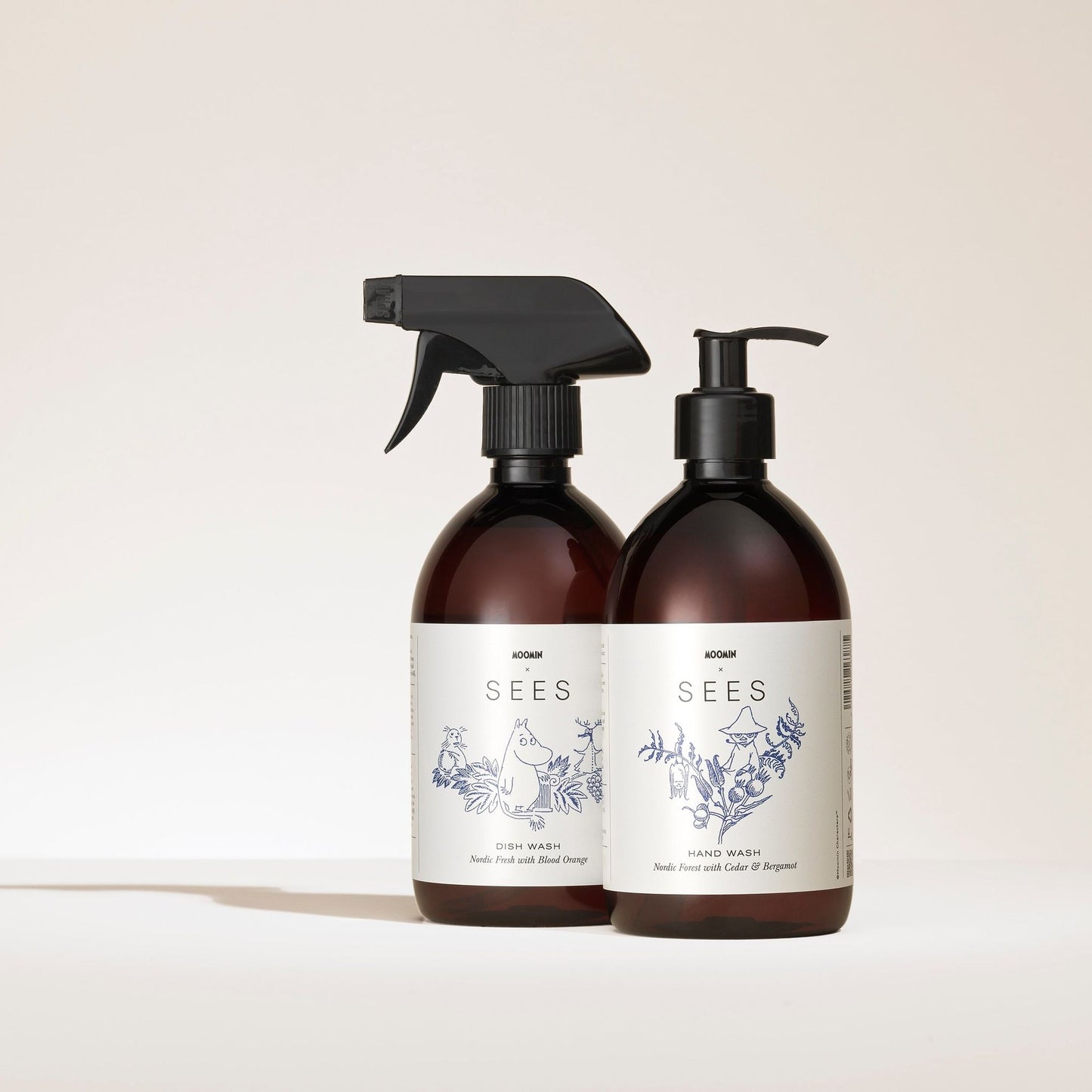Two brown plastic bottles from SEES Company with white labels featuring blue botanical illustrations; one spray labeled Dish Wash and one pump bottle labeled SEES x Moomin Hand Wash. Both are infused with essential oils.