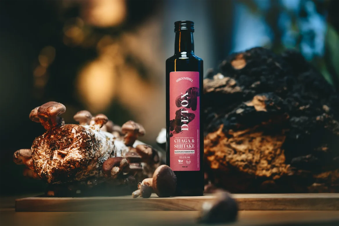 A bottle of Shroomwell Detox Elixir by Shroomwell rests on a wooden surface, surrounded by mushrooms and tree bark, highlighting the essence of a detox elixir powered by Chaga for natural detoxification.