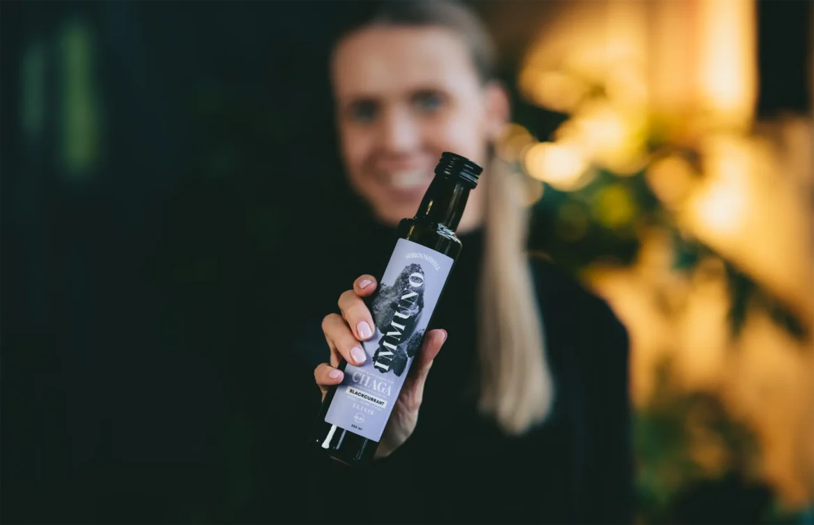 A blonde woman holds a dark glass bottle of Shroomwell Immuno Elixir Chaga & Blackcurrant—an immune-boosting supplement—toward the camera, set against a blurred green and yellow background.