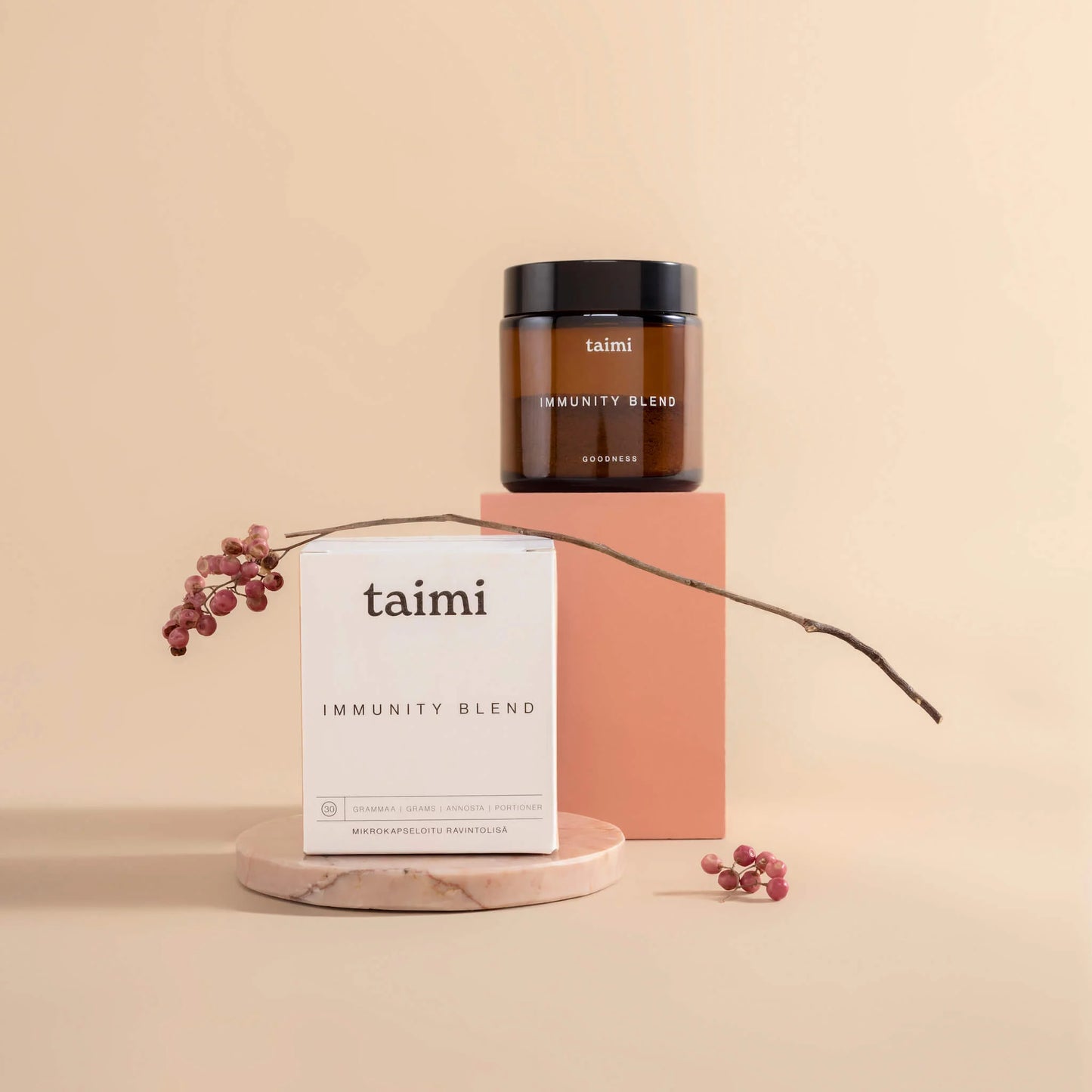 A jar and a box of Taimi Immunity Blend by Taimi are displayed on a marble platform alongside a pink block, with a branch of pink berries set against a minimalist beige background.