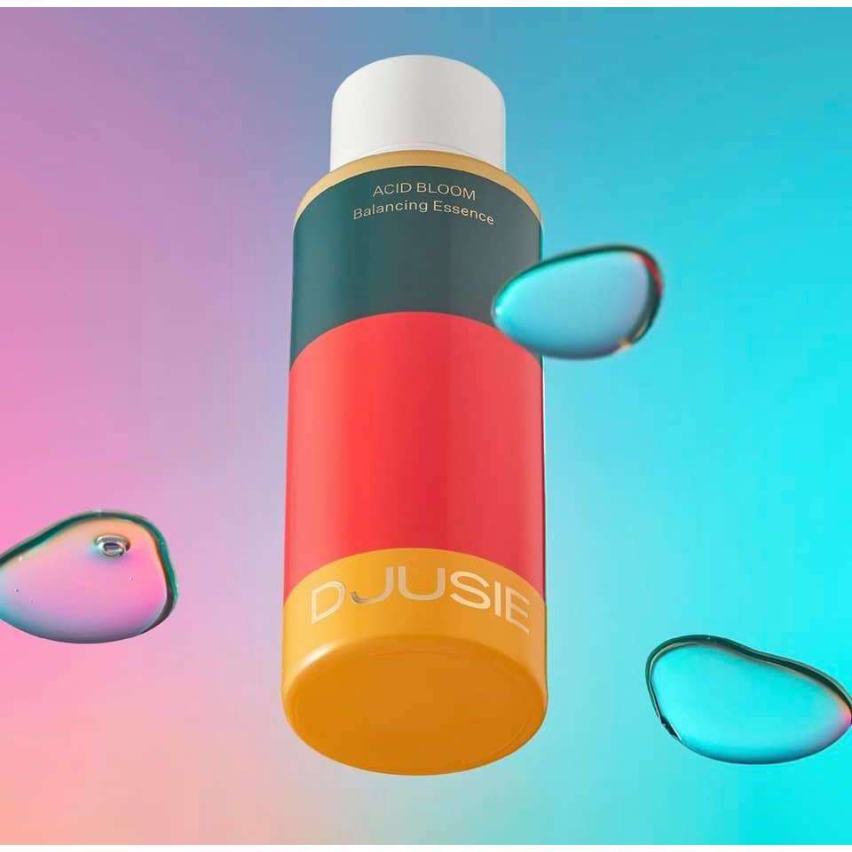 A vibrant bottle of Djusie Acid Bloom Balancing Essence floats against a pink and blue gradient background with three water droplets, showcasing Djusie’s brightening essence and its pH-balancing benefits.