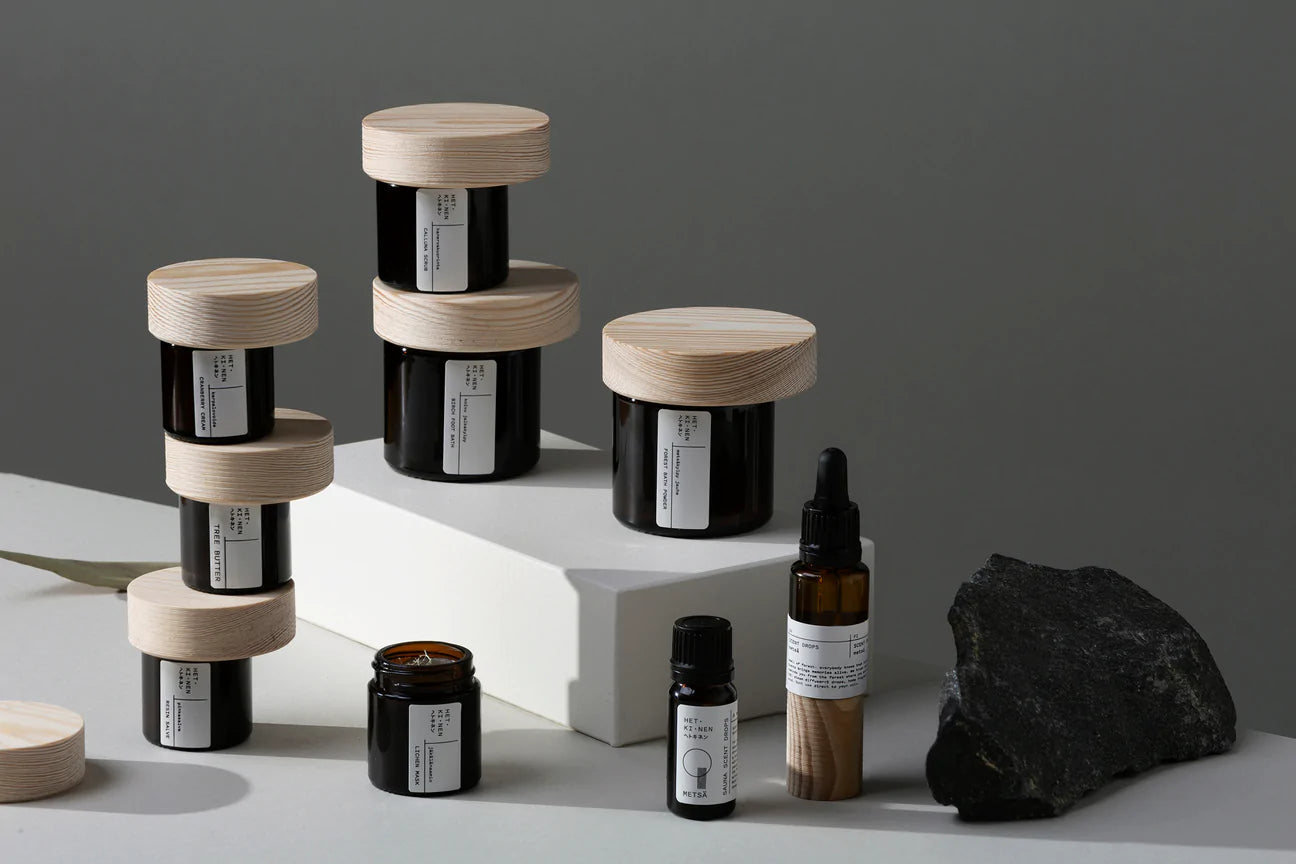 Hetkinen Metsä Calluna Scrub jars and bottles, with white labels, are elegantly arranged on white platforms beside a black stone, highlighting the natural skincare ritual of Hetkinen against a refined gray backdrop.