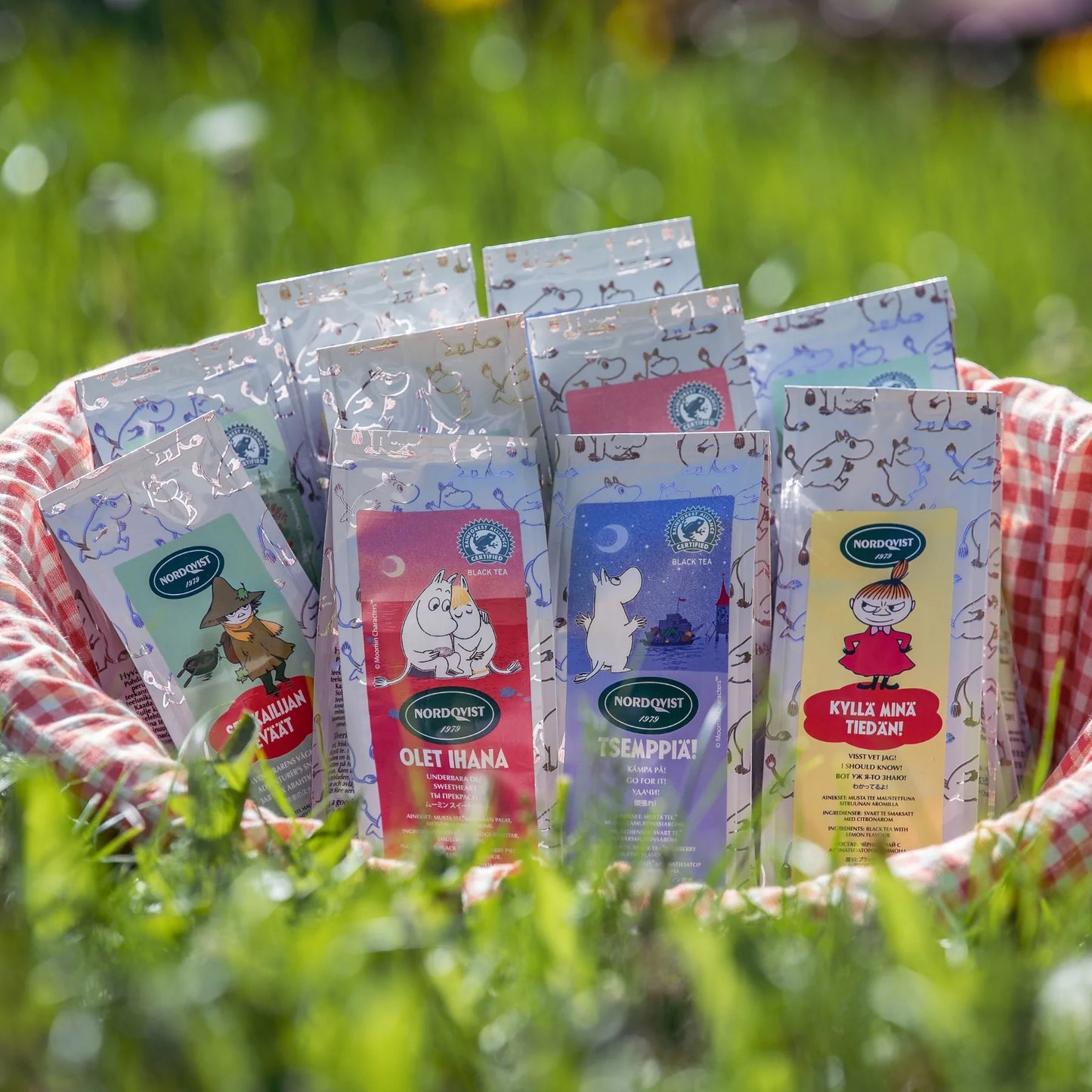 A basket with a red-and-white checkered cloth displays several Nordqvist Moomin Sweetheart tea packages—featuring Moomin characters and black tea with wild strawberry—outdoors on green grass in bright sunlight.