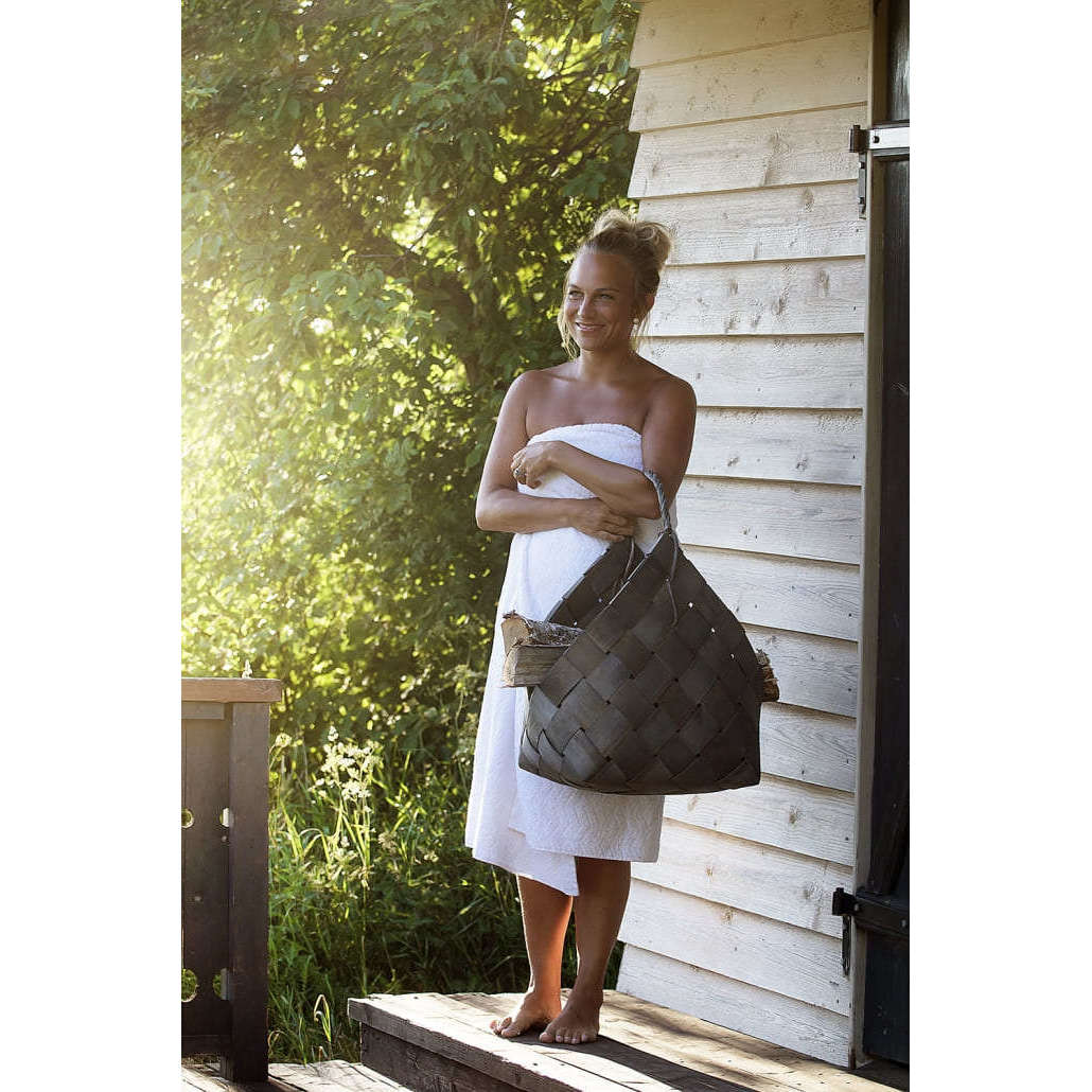 A woman in a white towel stands barefoot on a wooden porch, holding Emendo Relaxing Sauna Honey, with sunlight streaming in and green trees in the background.