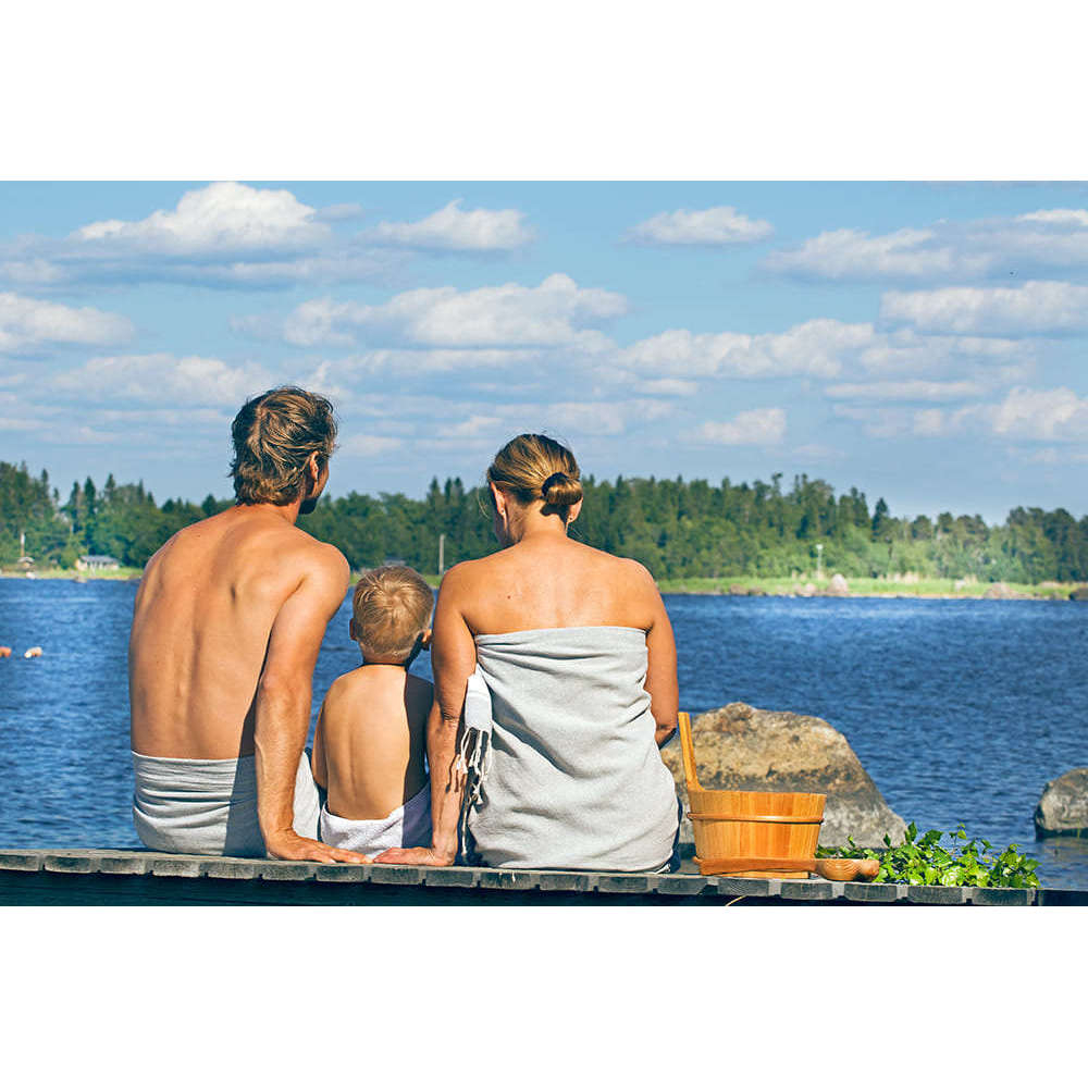 A man, woman, and child sit on a dock by a lake under a blue sky, wrapped in towels beside an Emendo Romantic Rose Sauna Honey jar, enjoying a serene retreat surrounded by trees and the moisturizing scent of roses.