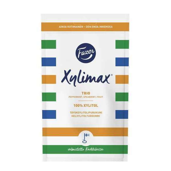 A white Fazer Xylimax Trio Full Xylitol Chewing Gum pack by Fazer features blue, green, orange, and yellow stripes. It promotes 100% xylitol for dental health and includes peppermint, spearmint, and fruit flavors.