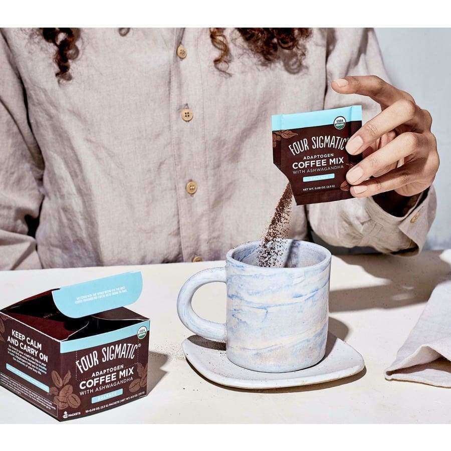 Someone pours Four Sigmatic Adaptogen Coffee Mix by Four Sigmatic into a white ceramic mug, while an open box of the coffee mix rests on the table nearby.