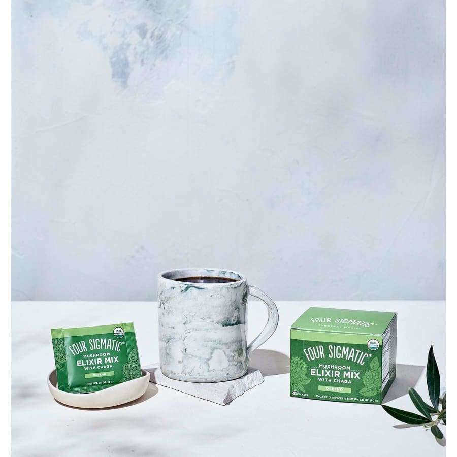 A marble mug on a coaster sits beside a small plate with a packet of Four Sigmatic Mushroom Elixir Mix—Chaga-powered for immune support—and a green box of the caffeine-free mix by Four Sigmatic, all on a light, minimal surface.