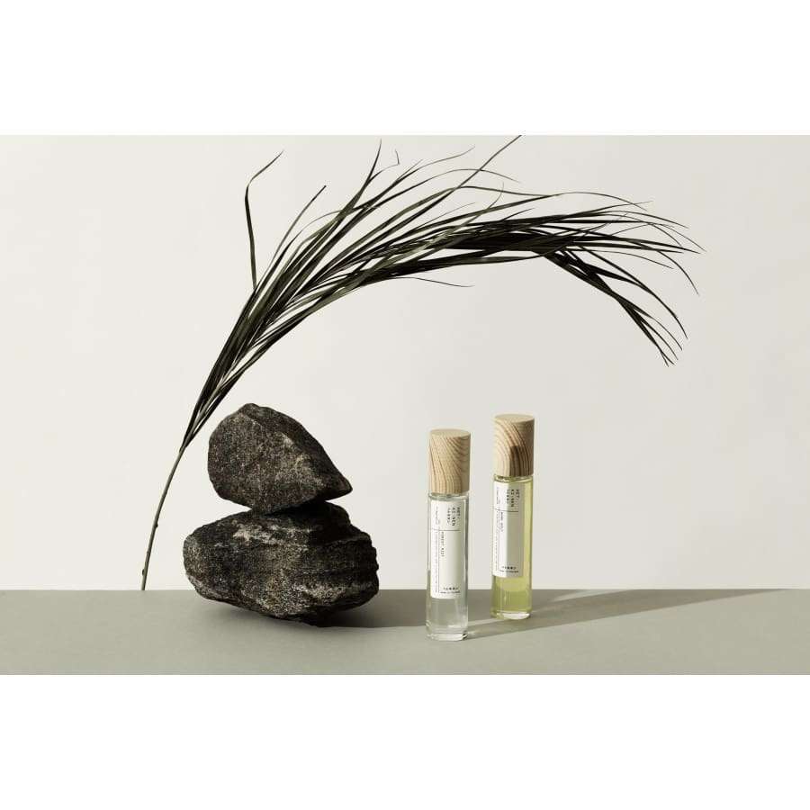 Two Hetkinen Fragrance Forest Riot bottles with wooden caps stand next to black stones and a curved dried palm leaf, forming a minimal, elegant display that highlights this vegan scent inspired by the natural forest.