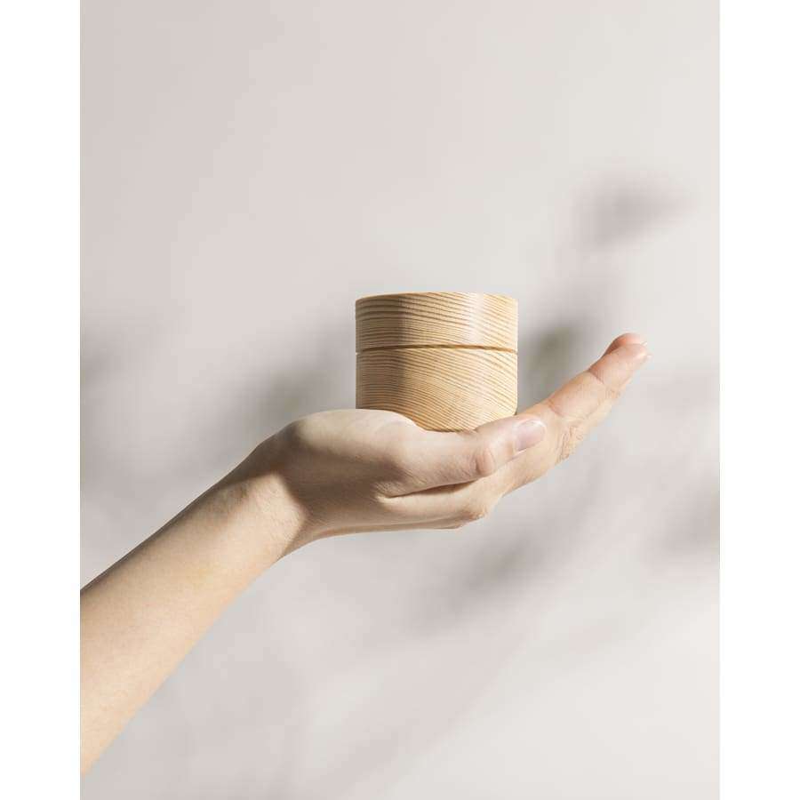 A hand holds a small, round, light wood container of Hetkinen Hand Balm Pine & Palmarosa against a plain, pale background with soft shadows.