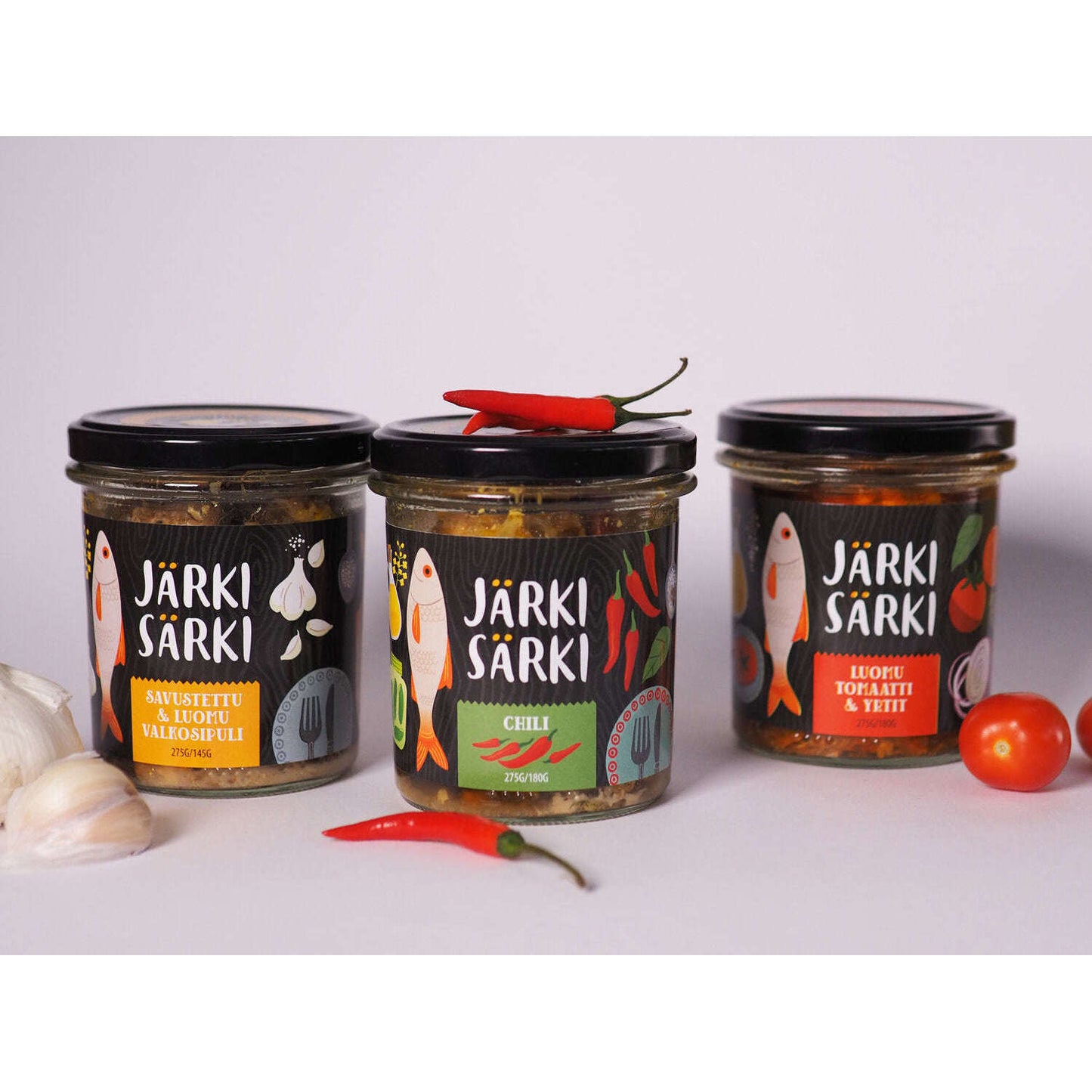 Three jars of Järki Särki Smoked with Organic Garlic fish preserves by Järki Särki, featuring vibrant labels, are shown alongside fresh garlic, red chili peppers, and a cherry tomato on a light background.