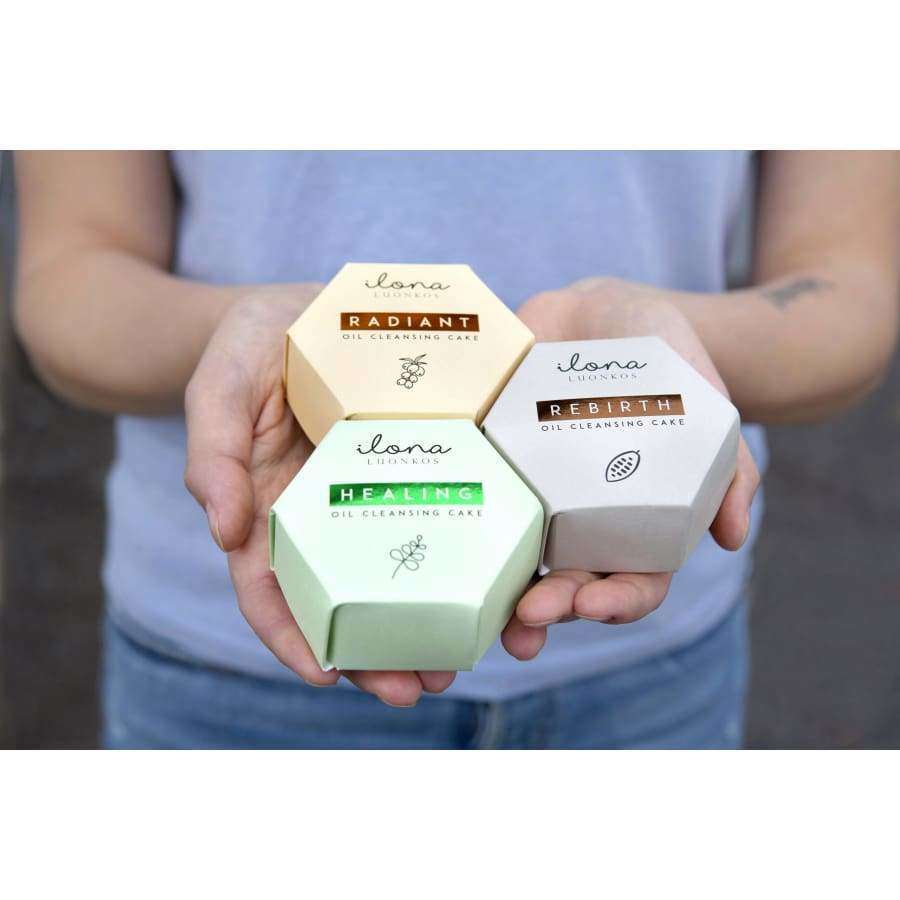 A person holds three hexagonal boxes labeled Radiant, Healing, and Rebirth by Luonkos. These vibrant packages contain the Luonkos Healing Oil Cleansing Cake, a natural facial cleanser for oily skin.