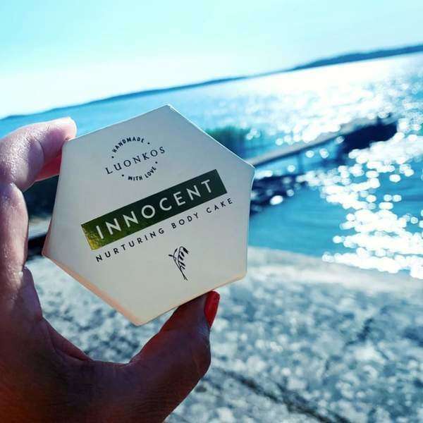 A hand holds a hexagonal Luonkos Innocent Nurturing Body Cake, crafted for dry and sensitive skin, near shimmering blue water with sunlight dancing on the surface.