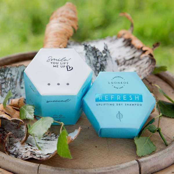Two hexagonal blue Luonkos Refresh Dry Shampoo packages with natural ingredients rest on wood among birch bark and green leaves. One reads “REFRESH Uplifting Dry Shampoo,” the other “Smiles! YOU LIFT ME UP.”.