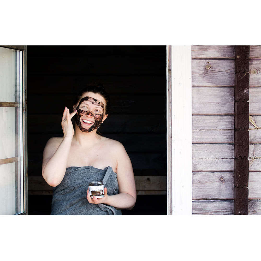 A person wrapped in a towel stands by an open window, smiling and applying Emendo Sauna Peat Mask with one hand while holding the Emendo jar in the other, next to a wooden wall.