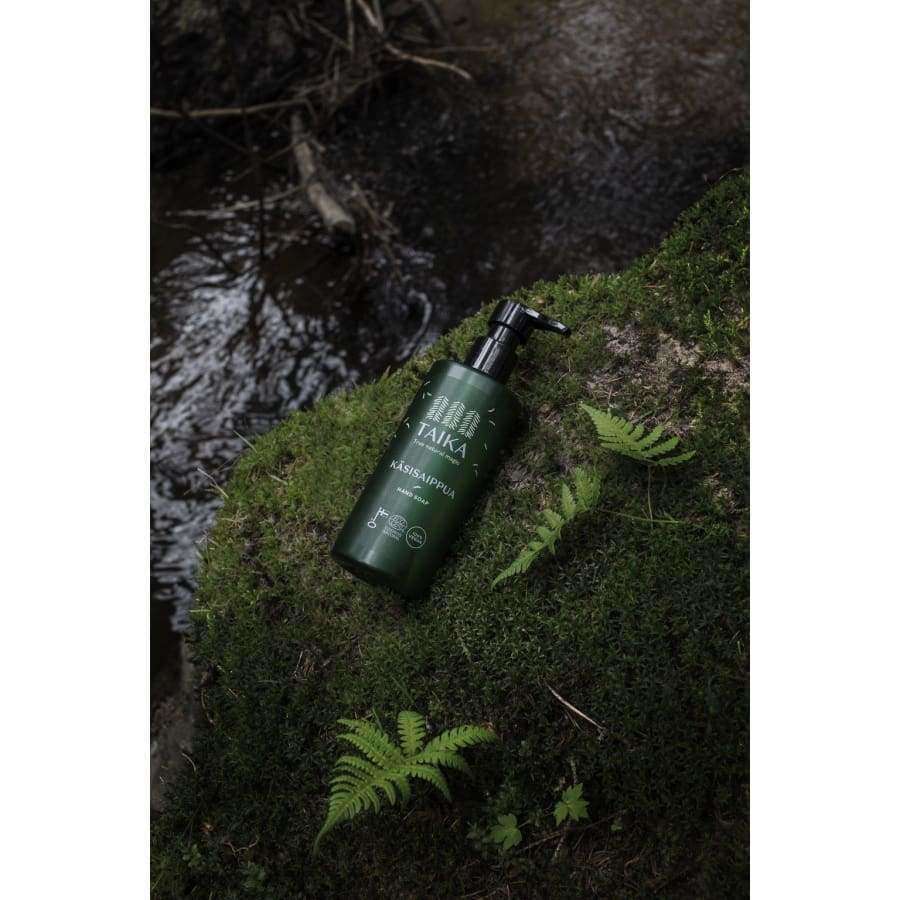A green bottle of Taika Liquid Hand Soap with a pump rests on moss near ferns and a stream, its forest setting evoking the refreshing scent of grapefruit and the clean purity of this vegan Taika product.