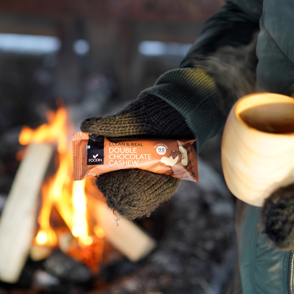 Eine Person mit dunklen Handschuhen hält einen Foodin Clean & Real Protein Bar Double Chocolate Cashew und einen Holzbecher vor einem Lagerfeuer mit brennenden Holzscheiten im Hintergrund.