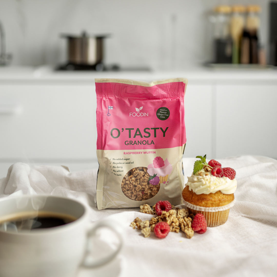 Eine Tüte Foodin O`Tasty Kauragranola Raspberry Muffin liegt auf einer weißen Fläche neben einem Kaffee, einem Himbeermuffin mit Sahne und Himbeeren und verstreuten Müslistücken. Im Hintergrund ist eine moderne Küche unscharf zu erkennen.
