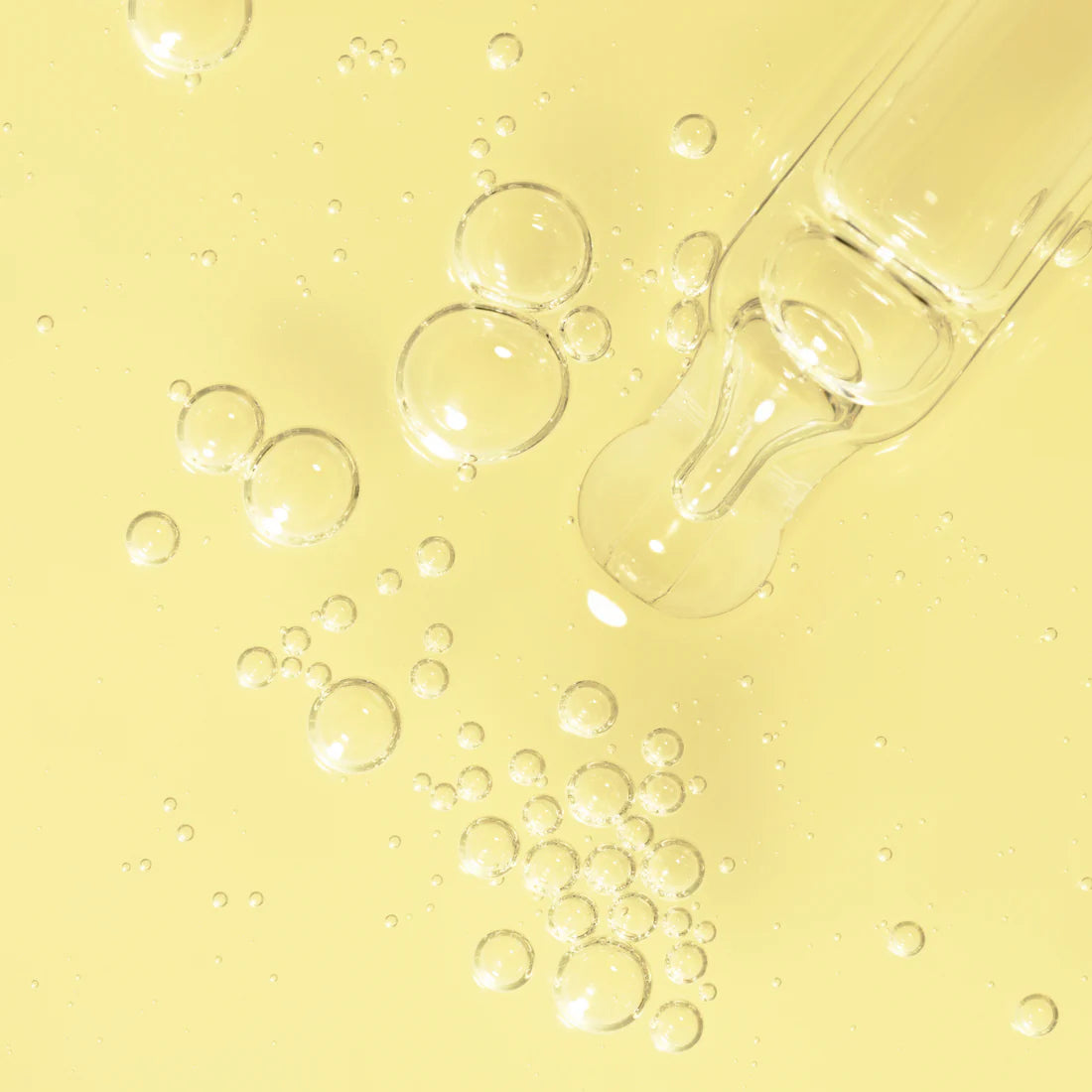 Ein klarer Glastropfer ruht auf einer blassgelben Oberfläche mit transparenten Bläschen, die die erfrischende Reinheit der Lumene Nordic-C Glow Boost Essence von Lumene und ihre strahlende Vitamin-C-Wirkung widerspiegeln.