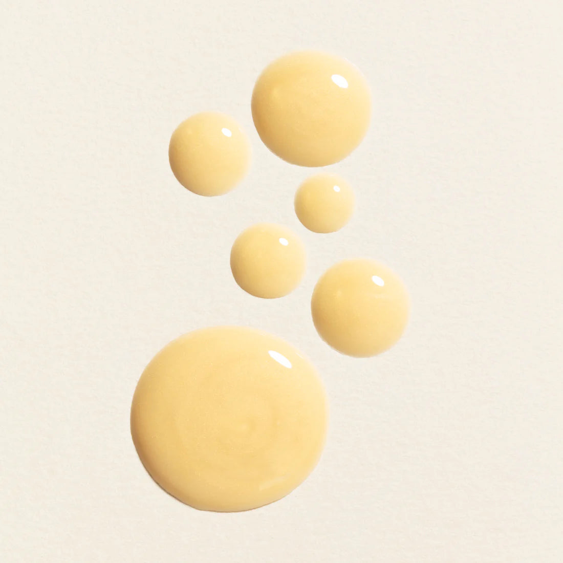 Sechs Tropfen des Lumene Nordic-C Glow Boost Eye Serum sind in einer lockeren, nach oben gerichteten Kurve auf einem hellen, strukturierten Hintergrund angeordnet. Die Tropfen sind unterschiedlich groß, wobei sich der größte unten und der kleinste oben befindet.