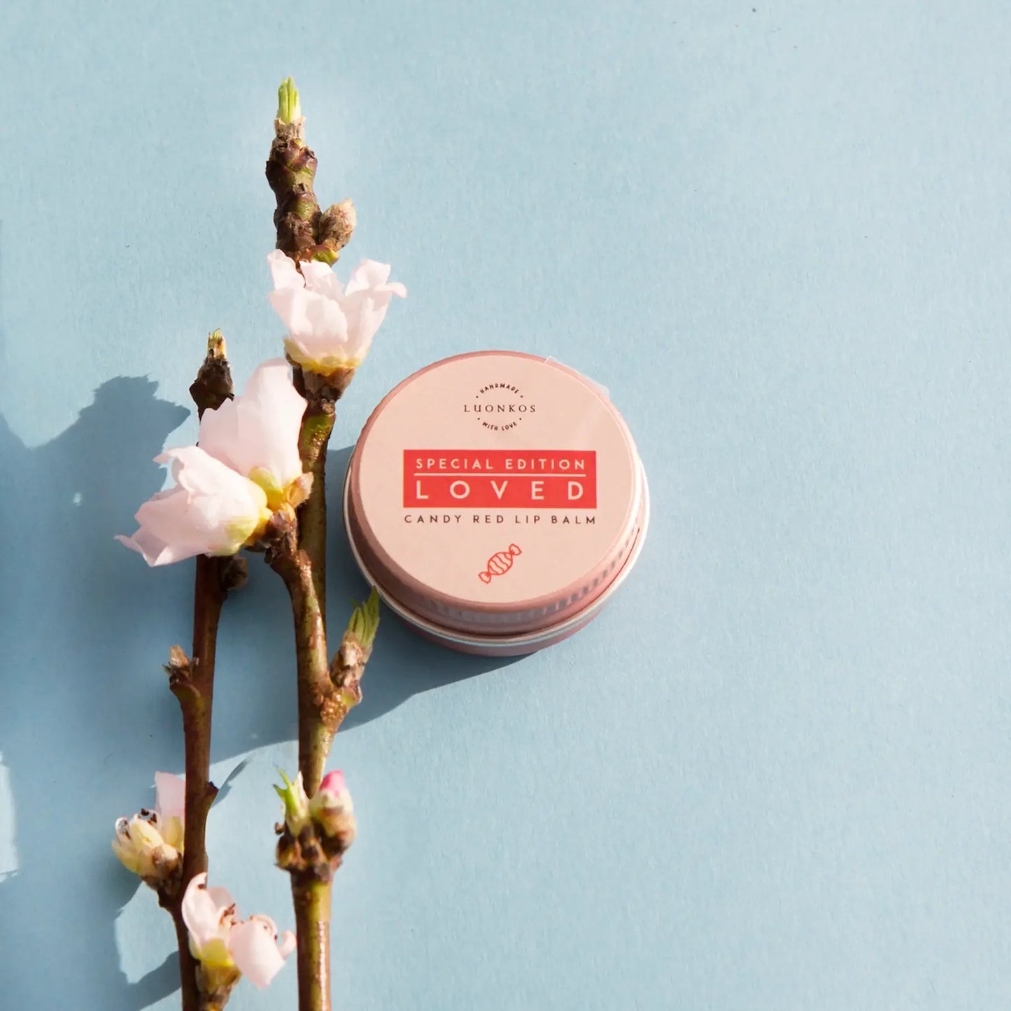 Eine Dose Luonkos Loved Special Edition Candy Red Lip Balm von Luonkos ruht auf einer hellblauen Fläche neben einem Zweig mit rosa Blüten.